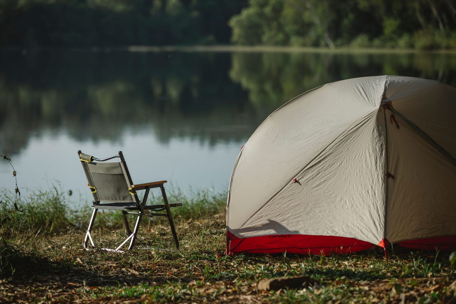 A folding camp chair and a tent on a grassy lakeshore in soft morning light